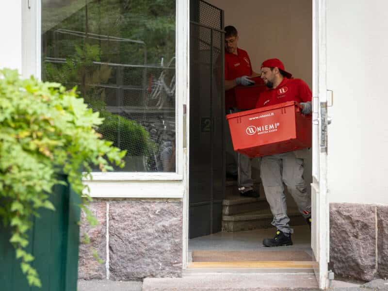 Muuttajat kantavat laatikoita kotimuutolla portaissa. Turvakengät ja työvaatteet näkyvät.